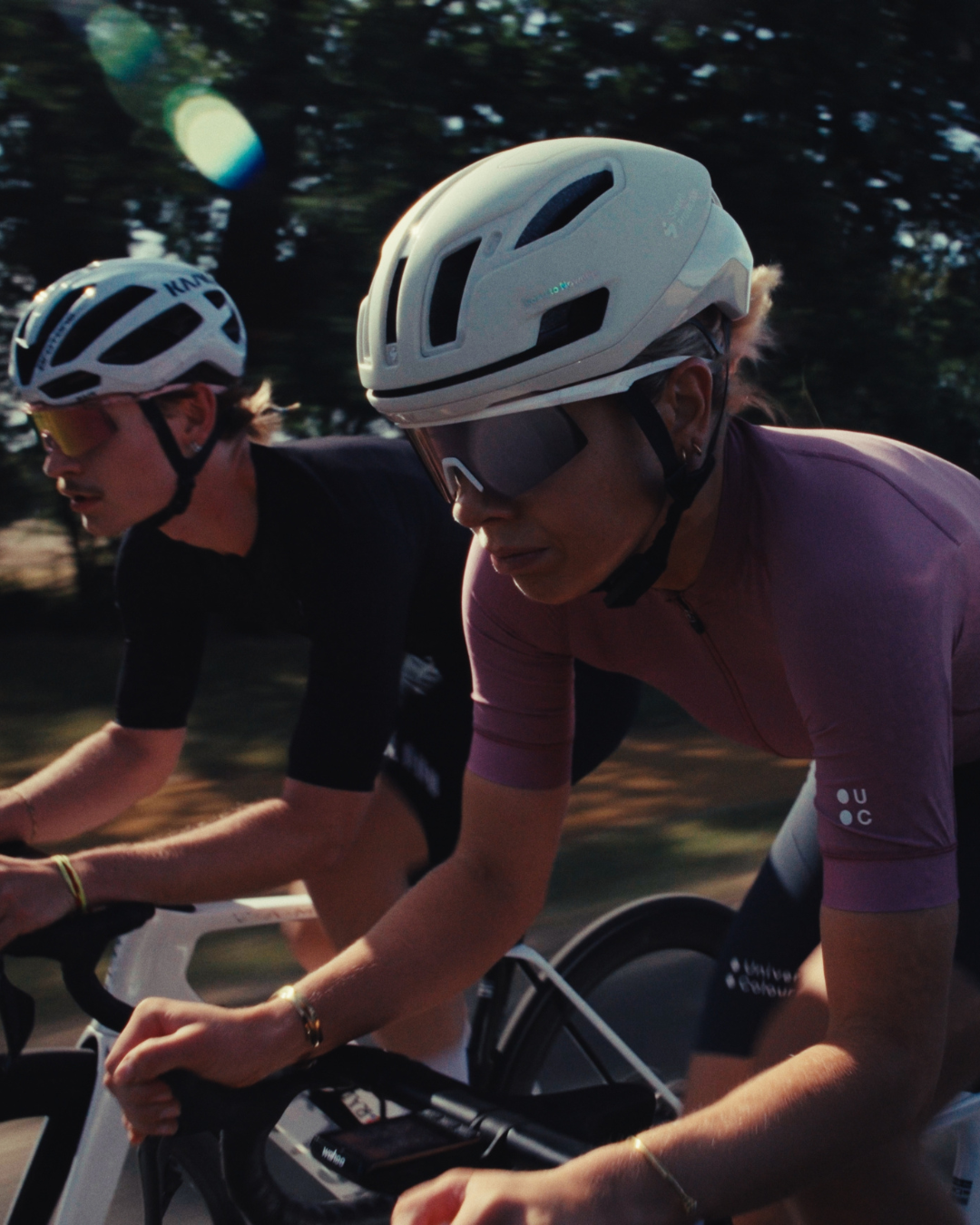 Two cyclists riding outdoors, with the foreground rider wearing a white cycling helmet, dark sunglasses, a mauve-pink short-sleeve cycling jersey, and black shorts. The background rider wears a black jersey and a white helmet.